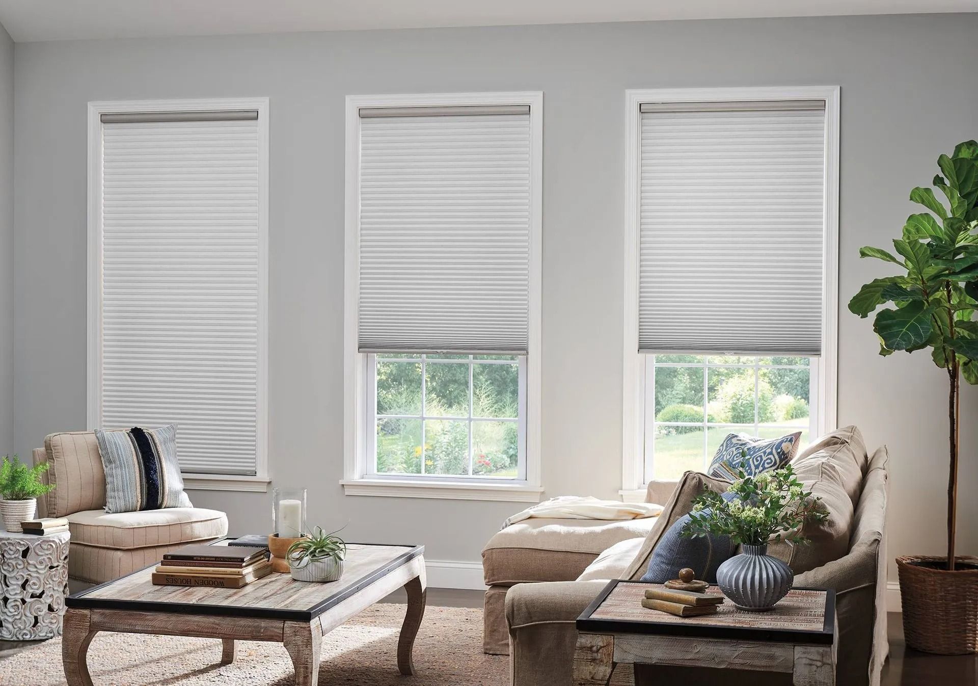 A bright living room features three windows with white pleated shades above two plush beige sofas and a wooden coffee table.
