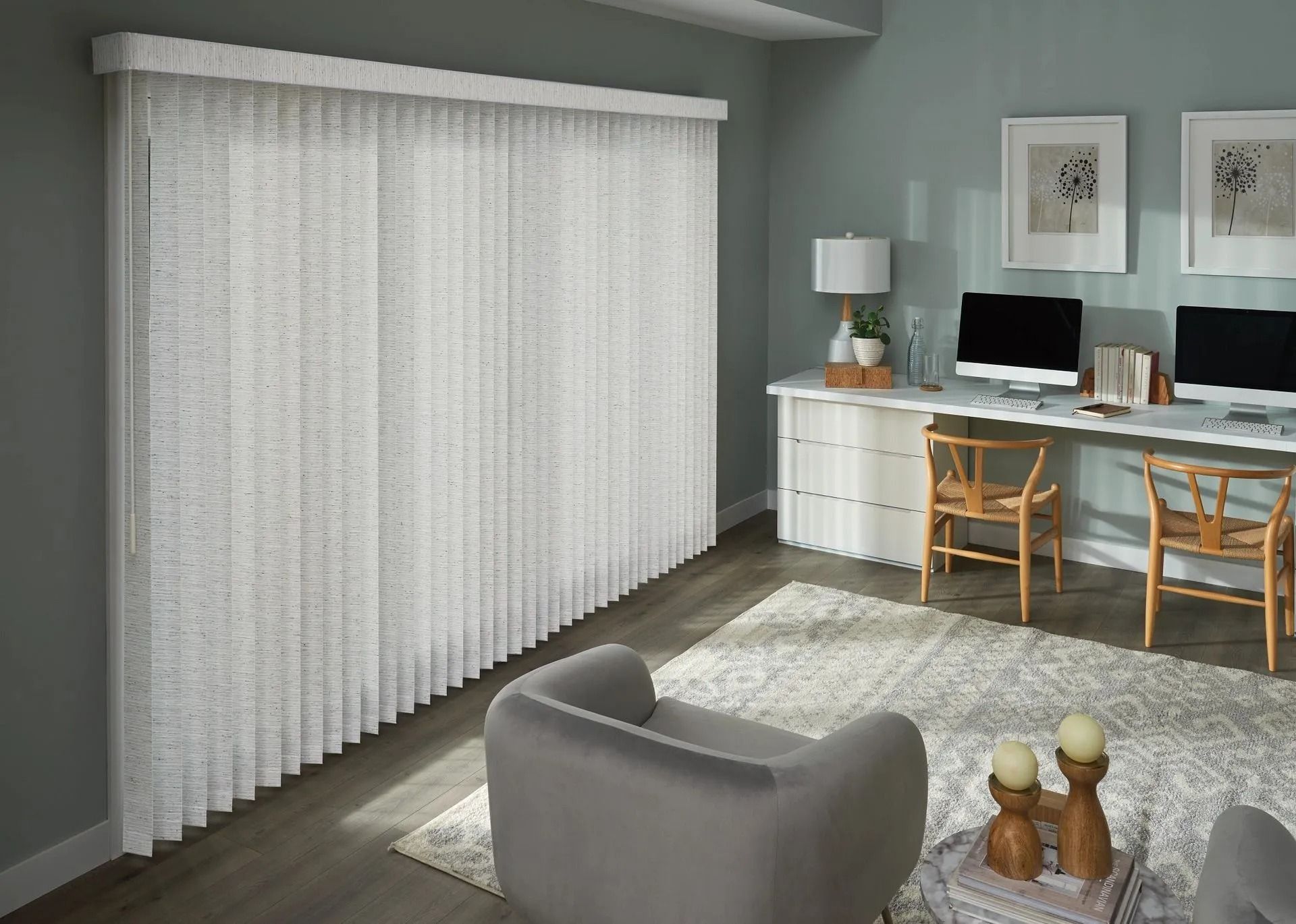 A modern home office with floor-to-ceiling white vertical blinds, a long desk with two computers, and a grey armchair.