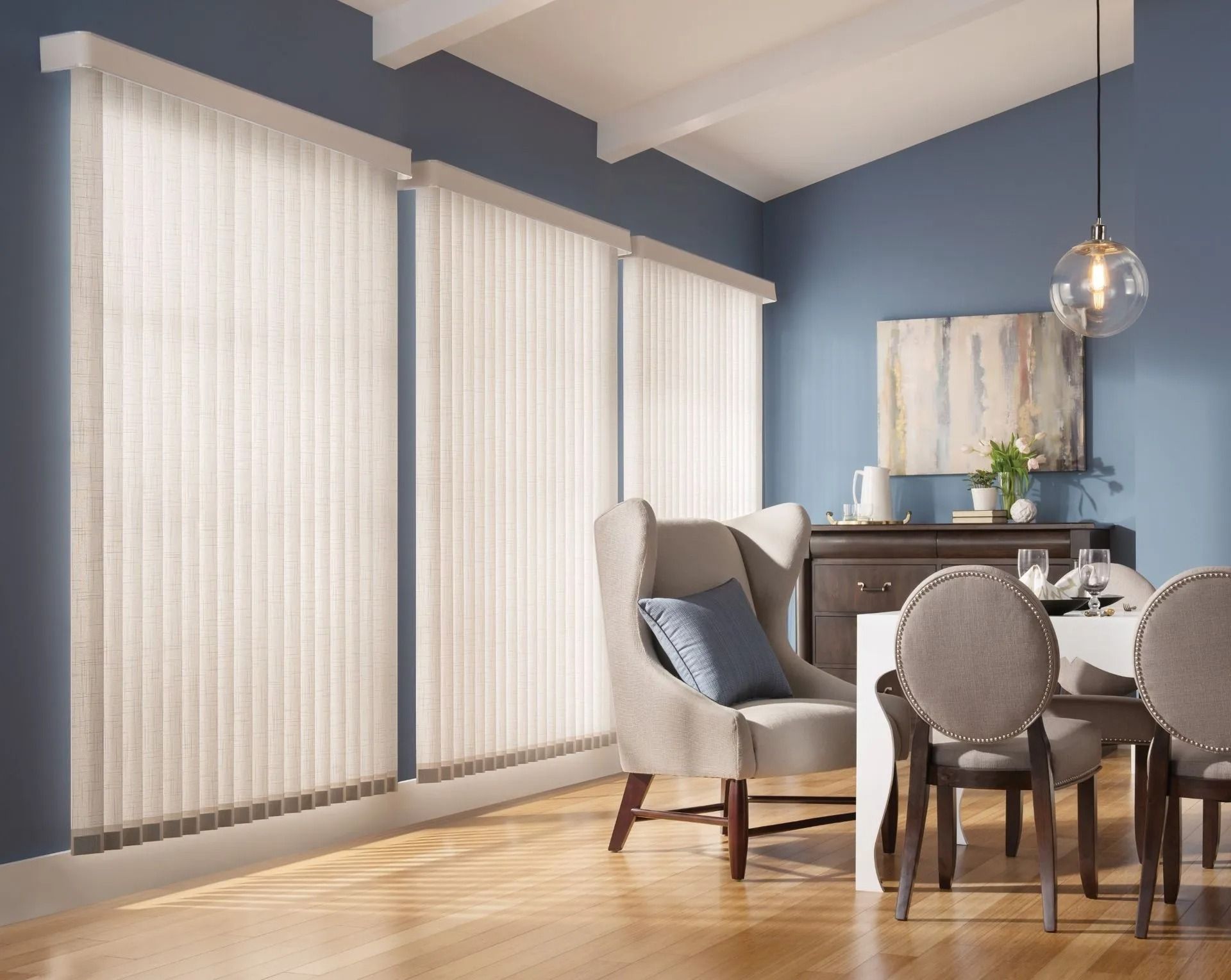 Modern dining room with vertical blinds on tall windows, a wingback chair, dining table with chairs, and blue walls.