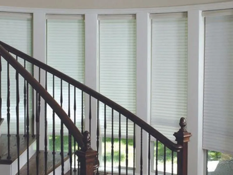 A curved staircase with dark wooden railings beside a row of windows covered by white pleated blinds.