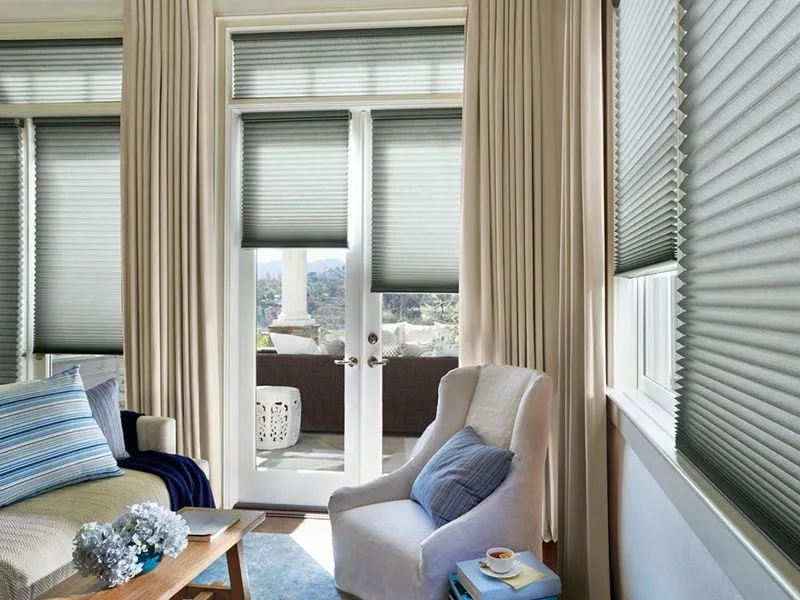 A bright living room with pleated shades on a glass door and windows, flanked by neutral curtains and furnished seating.