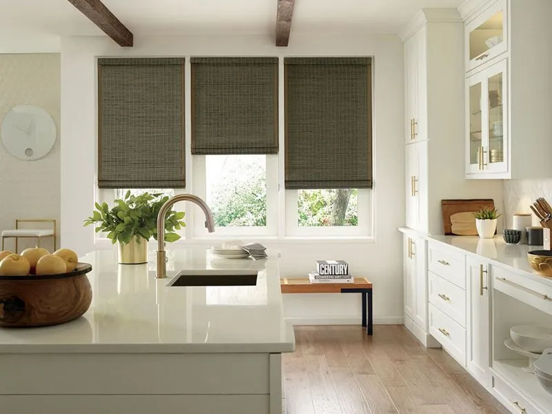A bright, modern kitchen featuring a white island, wooden ceiling beams, and three dark woven window shades.