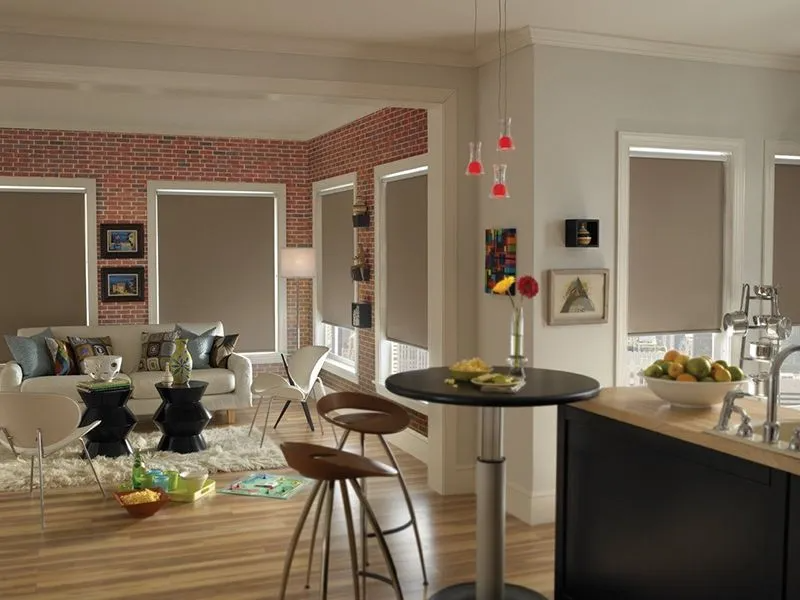 A modern, open-concept living area featuring neutral walls, a brick accent wall, light wood floors, and roller shades.