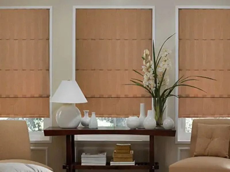 Three windows with tan roman shades above a dark wood table featuring a lamp, vase, flowers, and books between two chairs.