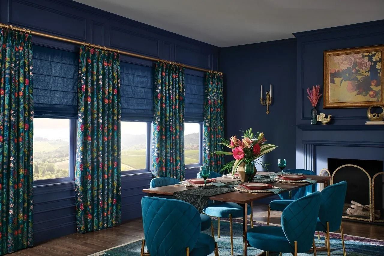 Dining room with deep blue walls, teal patterned curtains and Roman shades, a wooden table, and velvet teal chairs.