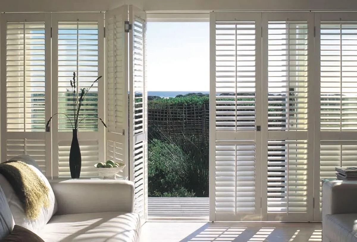 White louvered folding shutters frame a view of a lush green landscape and the sea through an open doorway.