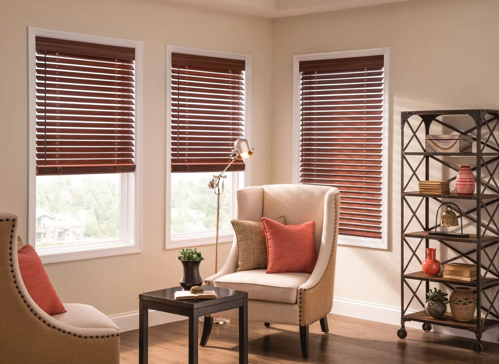 A cozy room features three windows with dark wood blinds, two upholstered chairs with orange pillows, and a metal shelf.