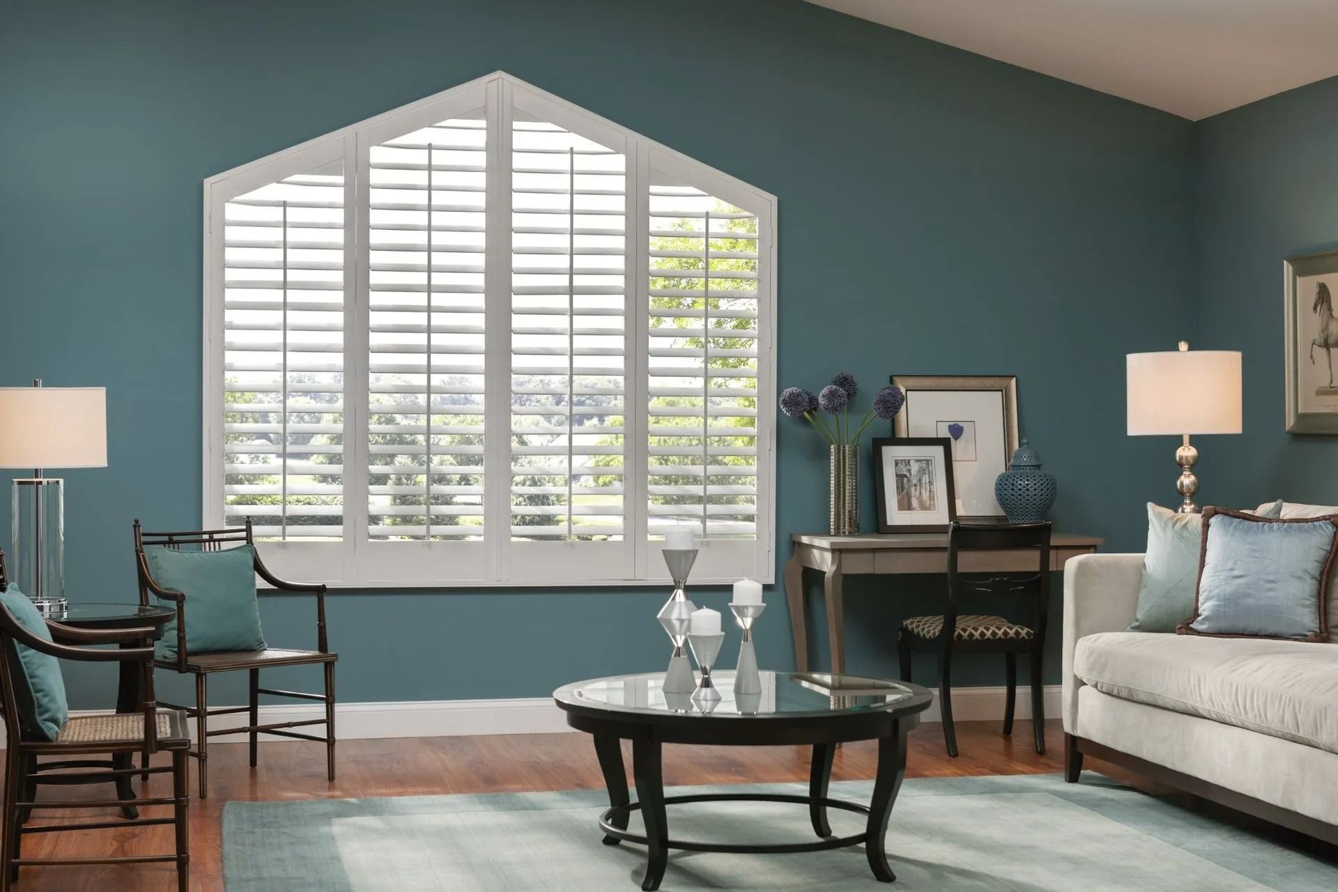 A living room with teal walls, a white pentagonal window with shutters, a round coffee table, and a neutral sofa.