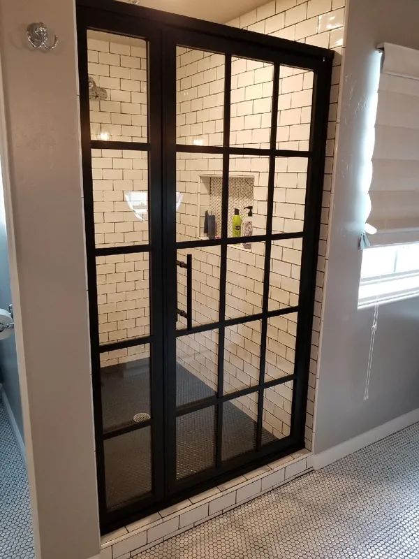 Black-framed glass shower door with grid design, white subway tile walls. Dark floor, beige countertop, and white blinds.