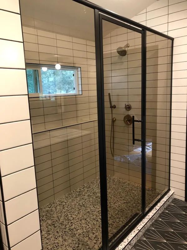 Modern shower with black-framed glass doors, beige tiled walls, pebble floor, and black and white patterned floor outside.
