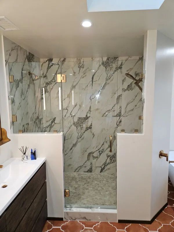 Bathroom with marble shower, glass doors, gold hardware, terracotta floor, and white walls.