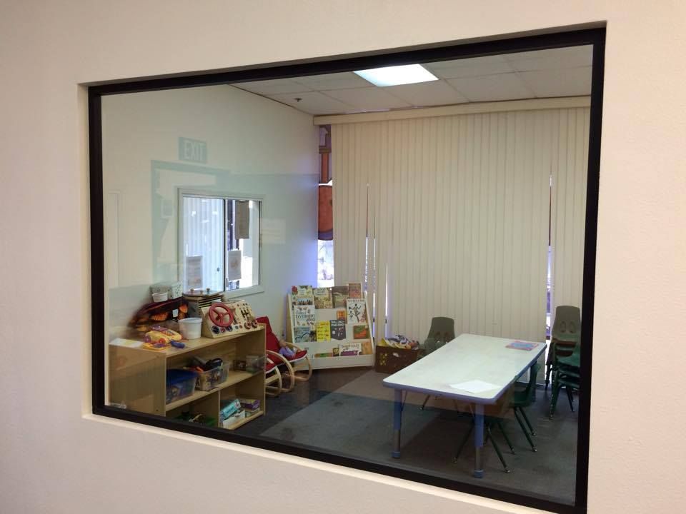 Room with child-sized furniture viewed through a window: table, chairs, toy shelves, blinds on a wall.