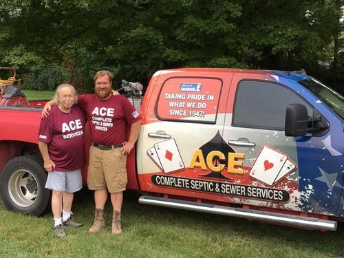 Two people standing beside a red ACE septic service truck in a wooded outdoor setting