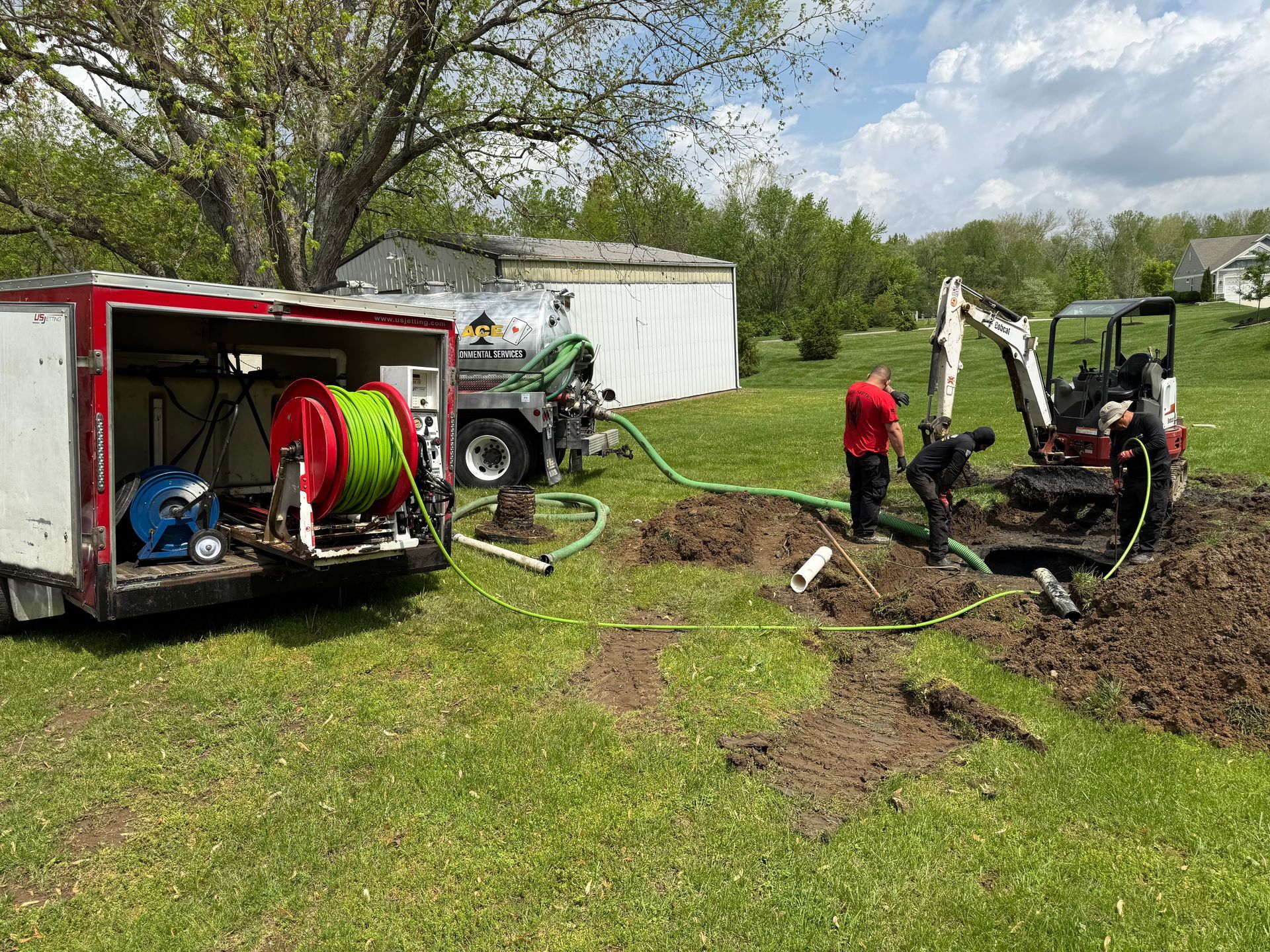 Workers using a mini excavator and hose reel near a utility truck in a muddy yard