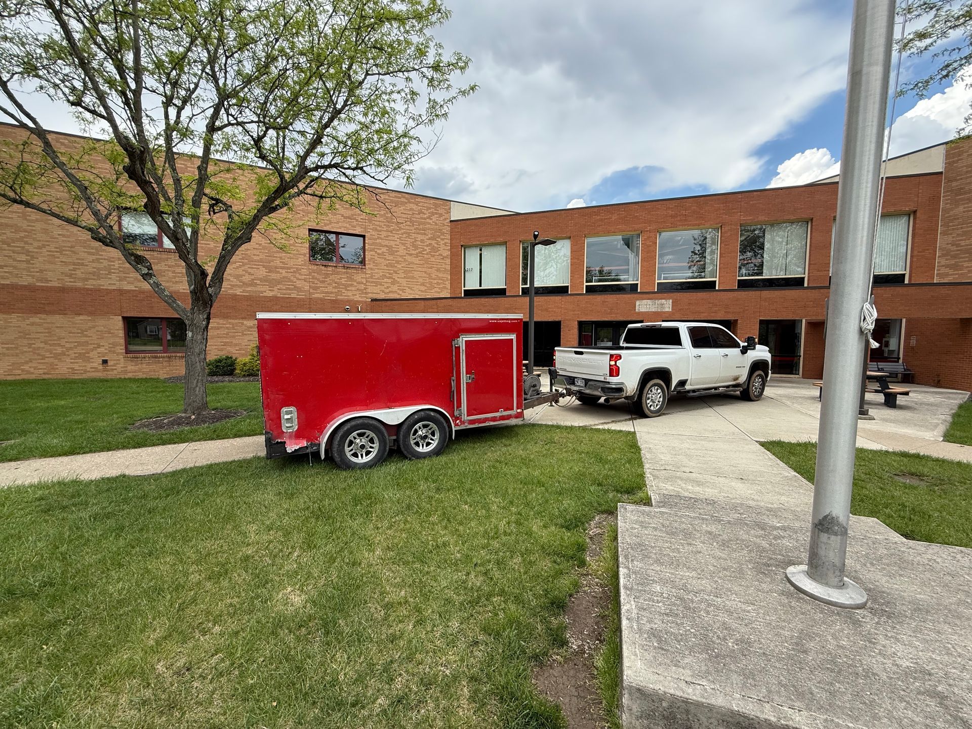 Red trailer and white pickup truck outside a brick building with a lawn and flagpole.