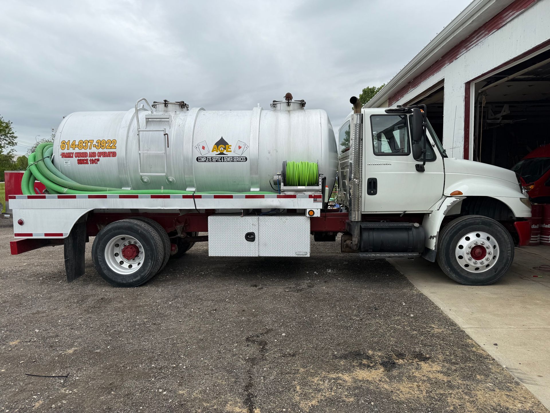 White vacuum truck parked beside a garage, with a silver tank and red stripes on the side.