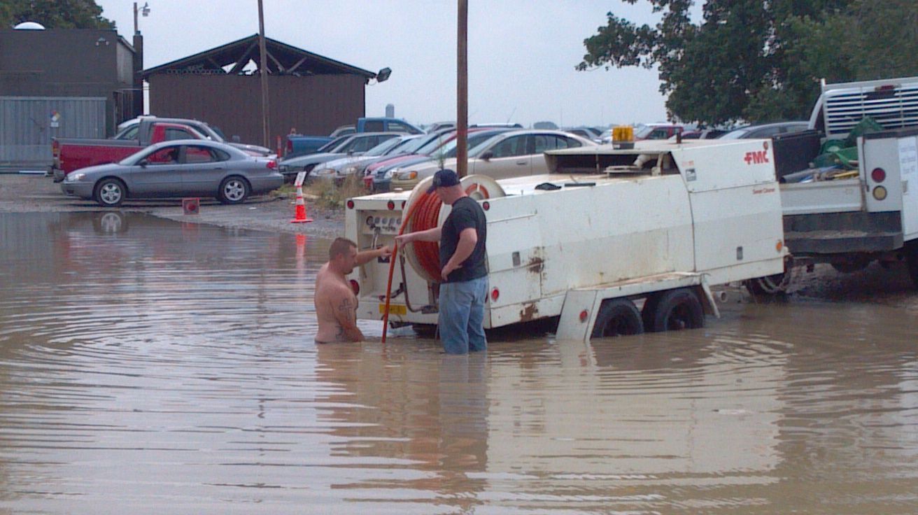 Workers stand in floodwater beside a white utility truck in a flooded parking lot.