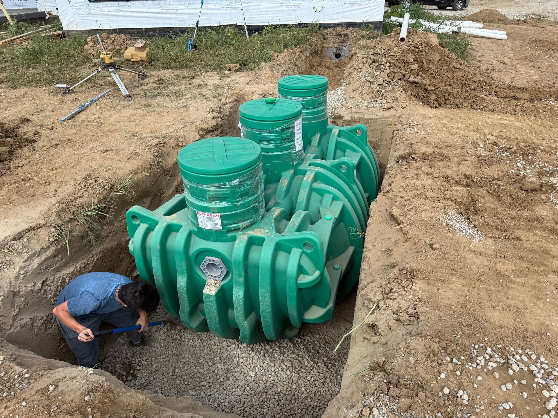 Worker inspecting a large green septic tank installation in a dirt excavation site