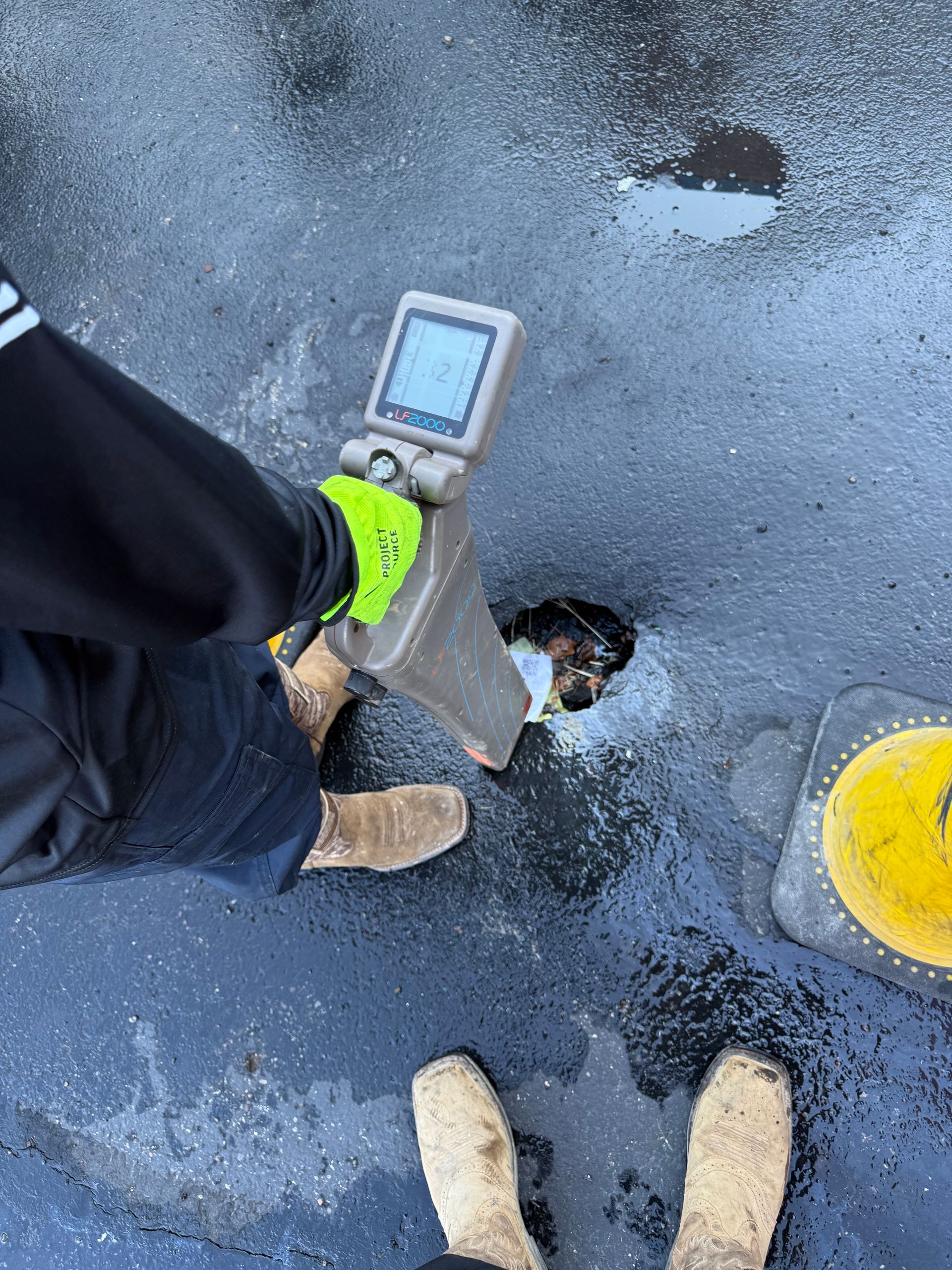 Person using a handheld meter to inspect a wet asphalt surface with potholes and a yellow bucket nearby