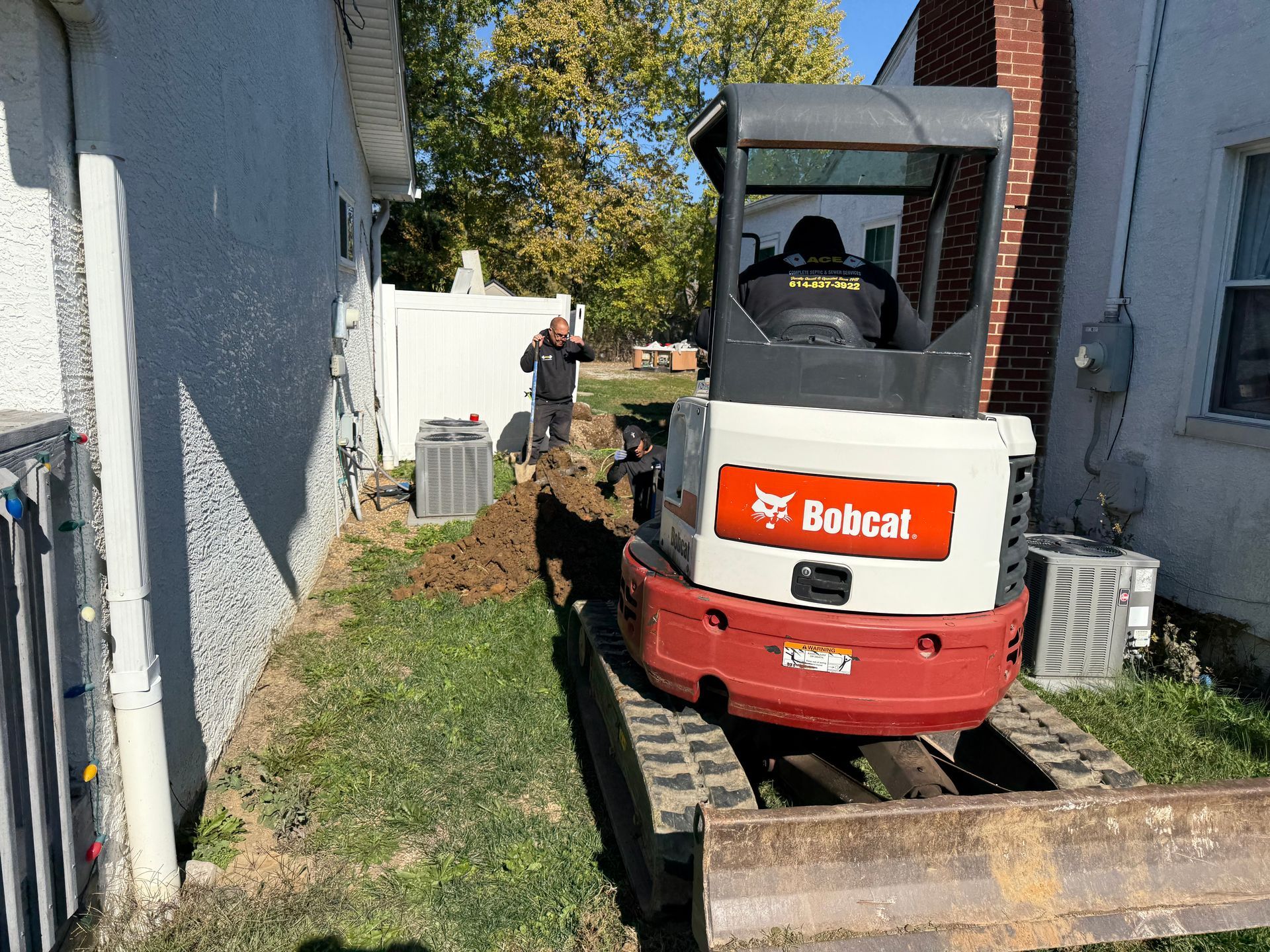Red Bobcat excavator on a narrow path between buildings, with a person working near the back.