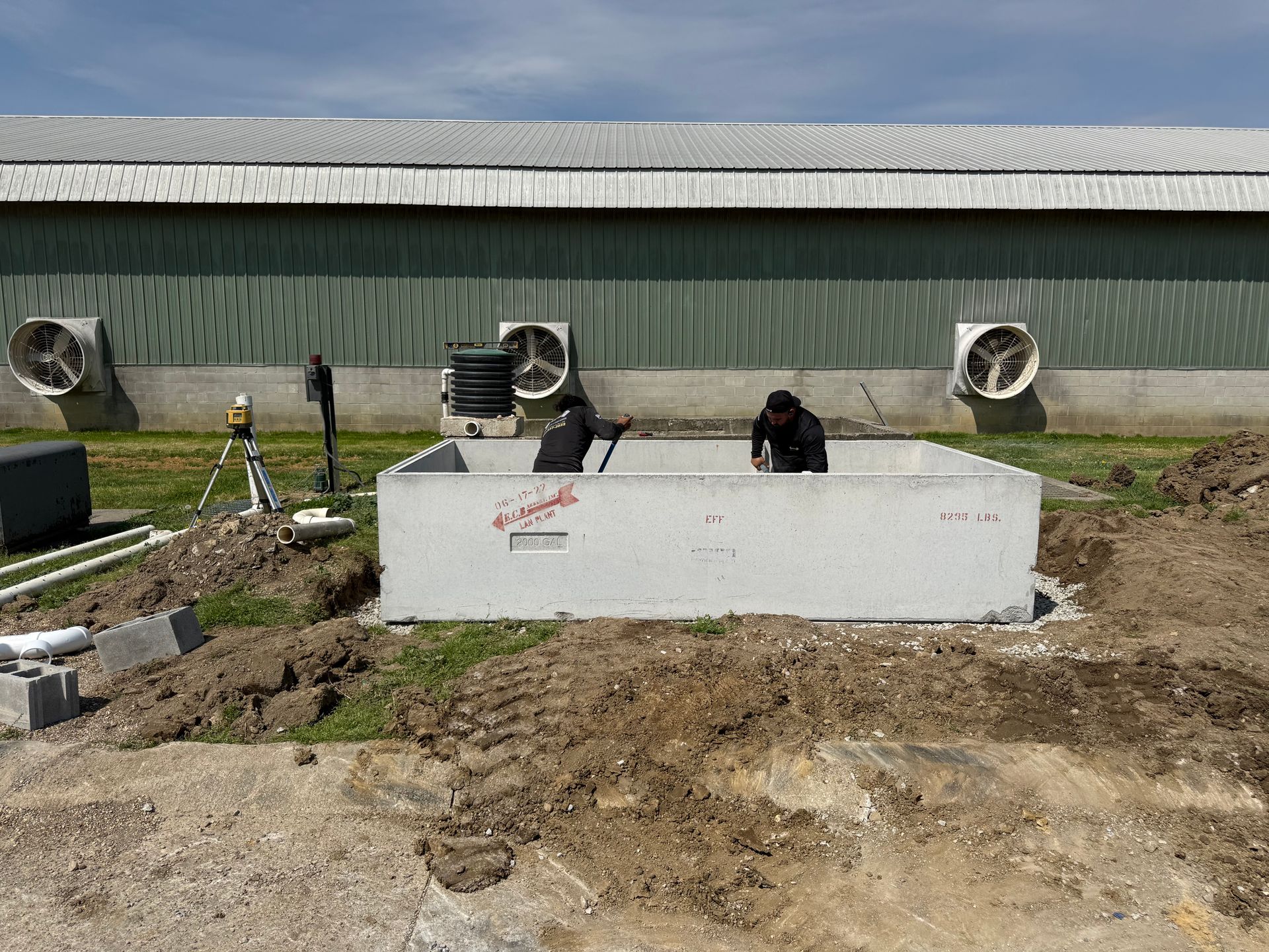 Workers installing a concrete barrier in a muddy field beside a long metal building