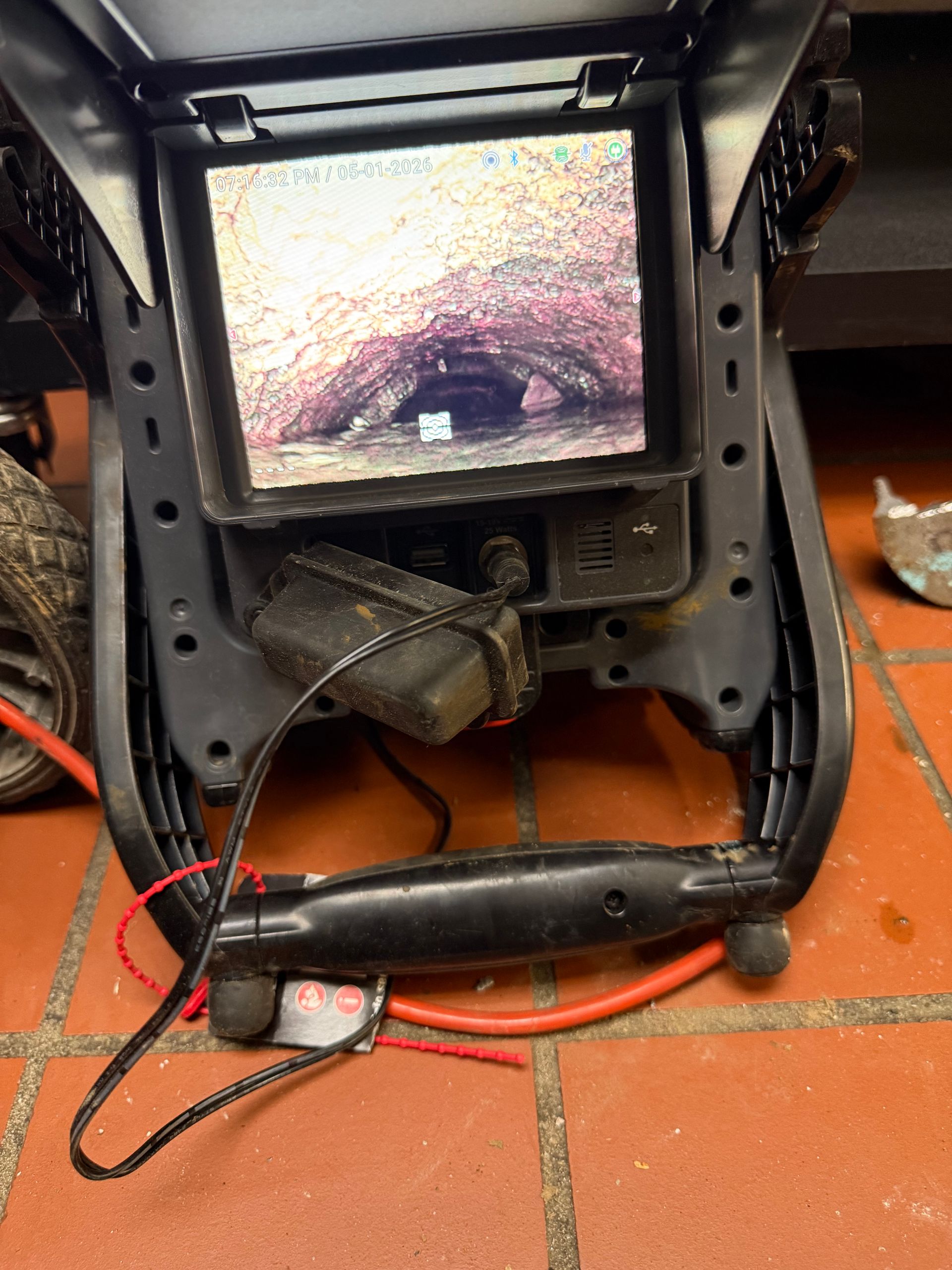 Black-framed portable screen or device mounted on orange tile floor with cables and a plug attached