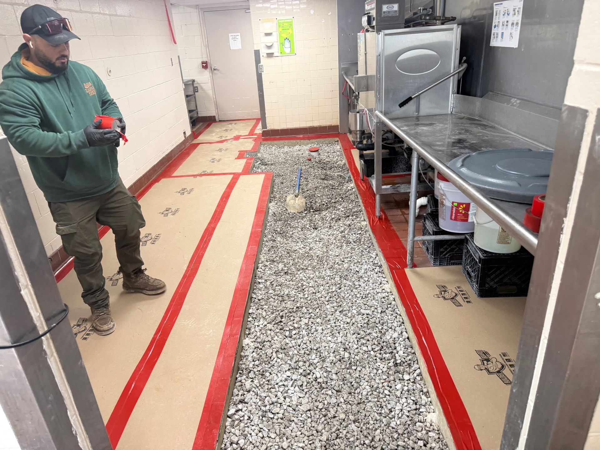 Worker standing in a narrow kitchen aisle with red floor tape and exposed subfloor under repair