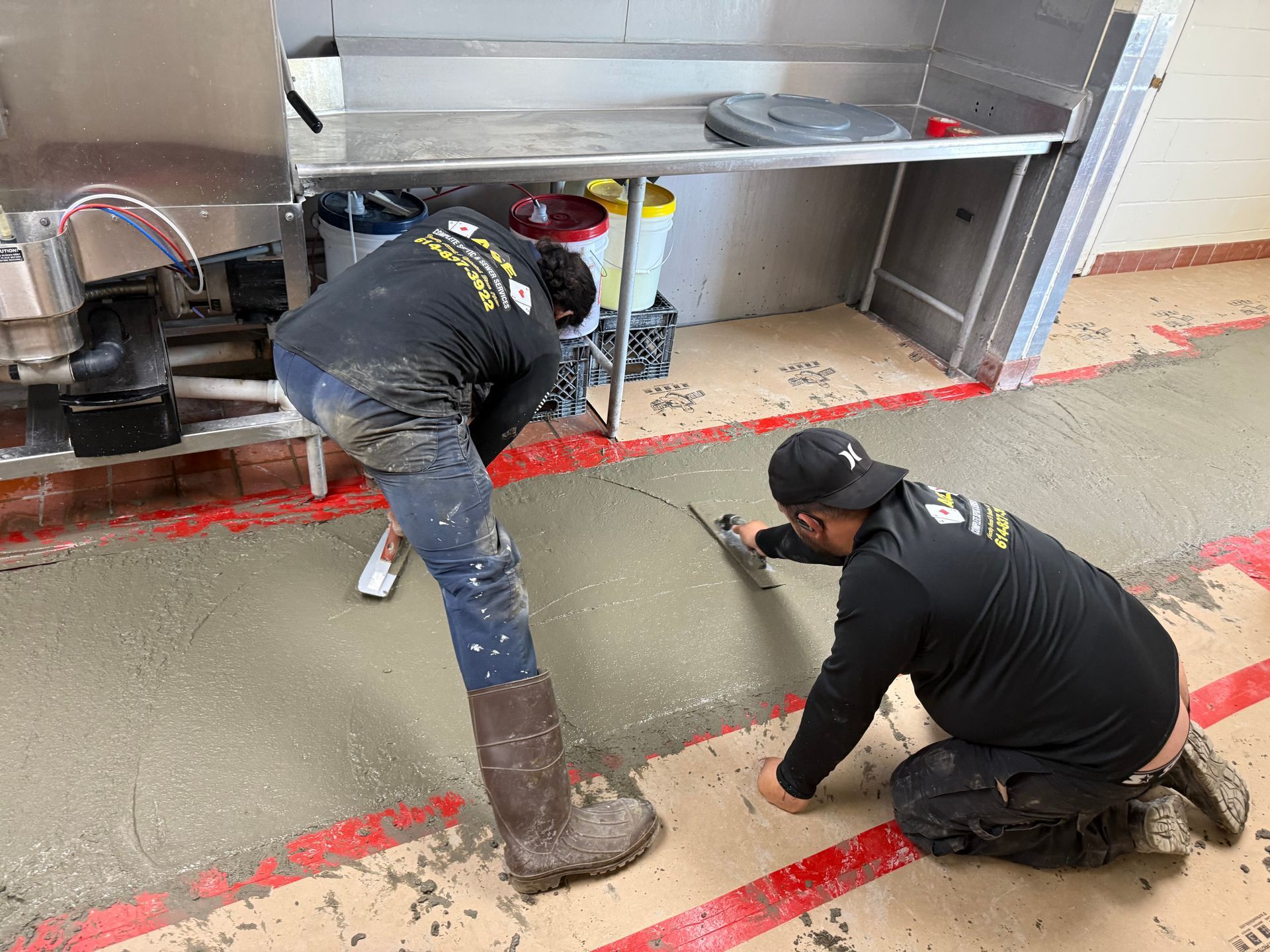 Two workers on a concrete floor, one kneeling and one bent over, working near red tape in a kitchen area