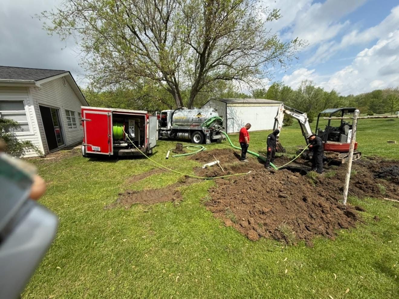 Workers with a small excavator and truck digging a yard beside a house under a cloudy sky