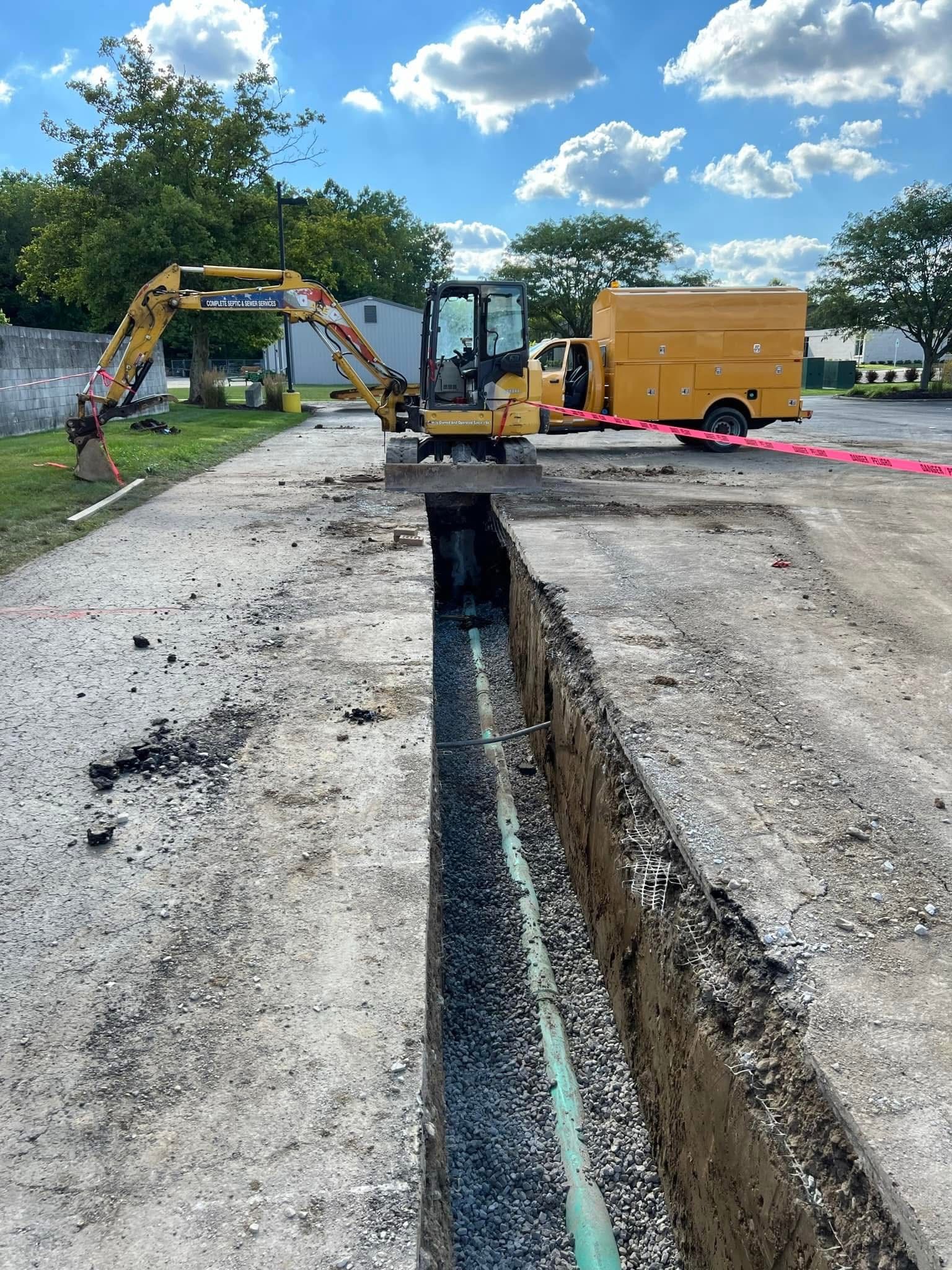 Construction site with excavator digging a long trench beside a gravel road under a blue sky