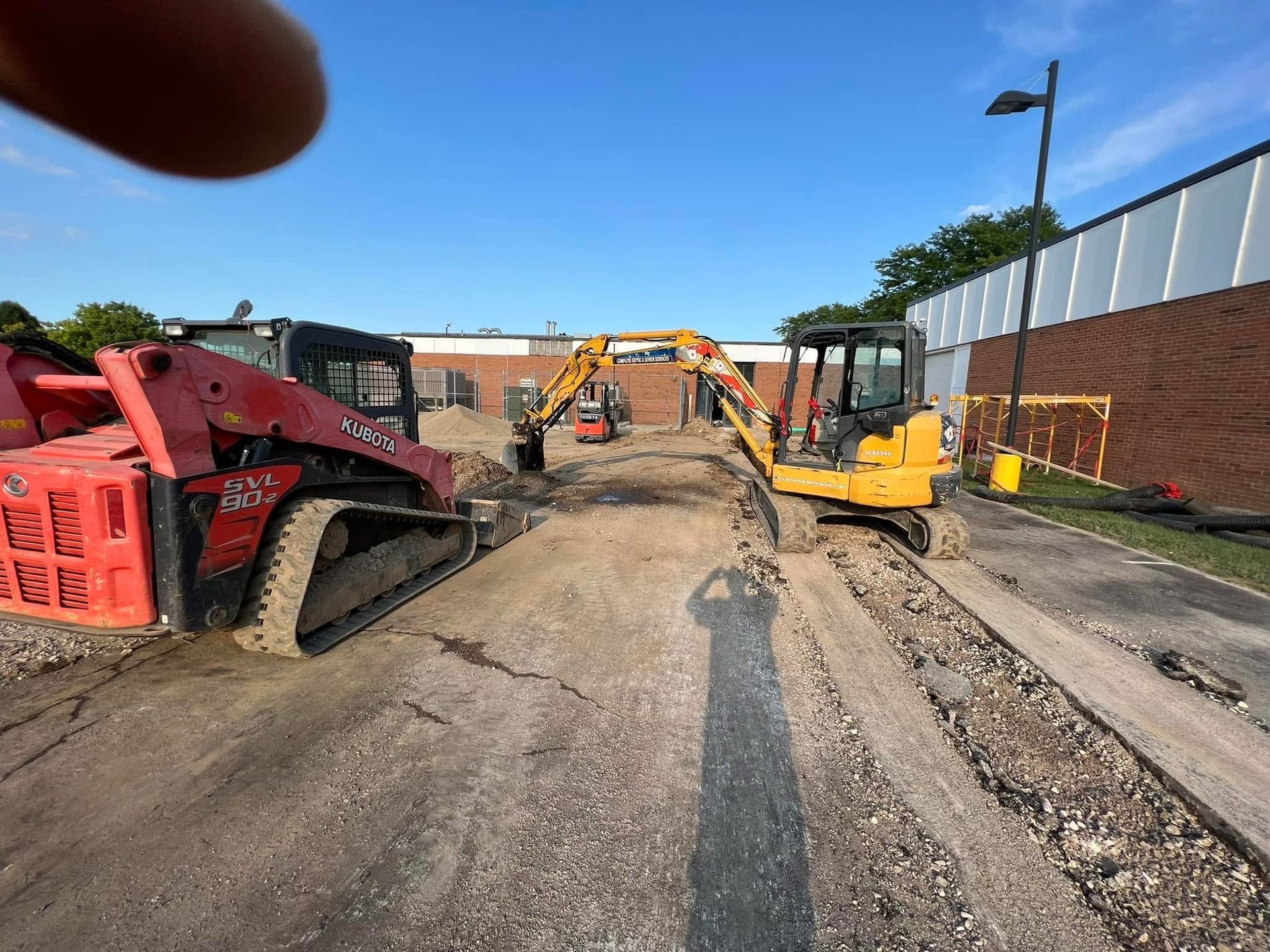 Construction site with excavators and skid steers grading a muddy dirt road beside a brick wall