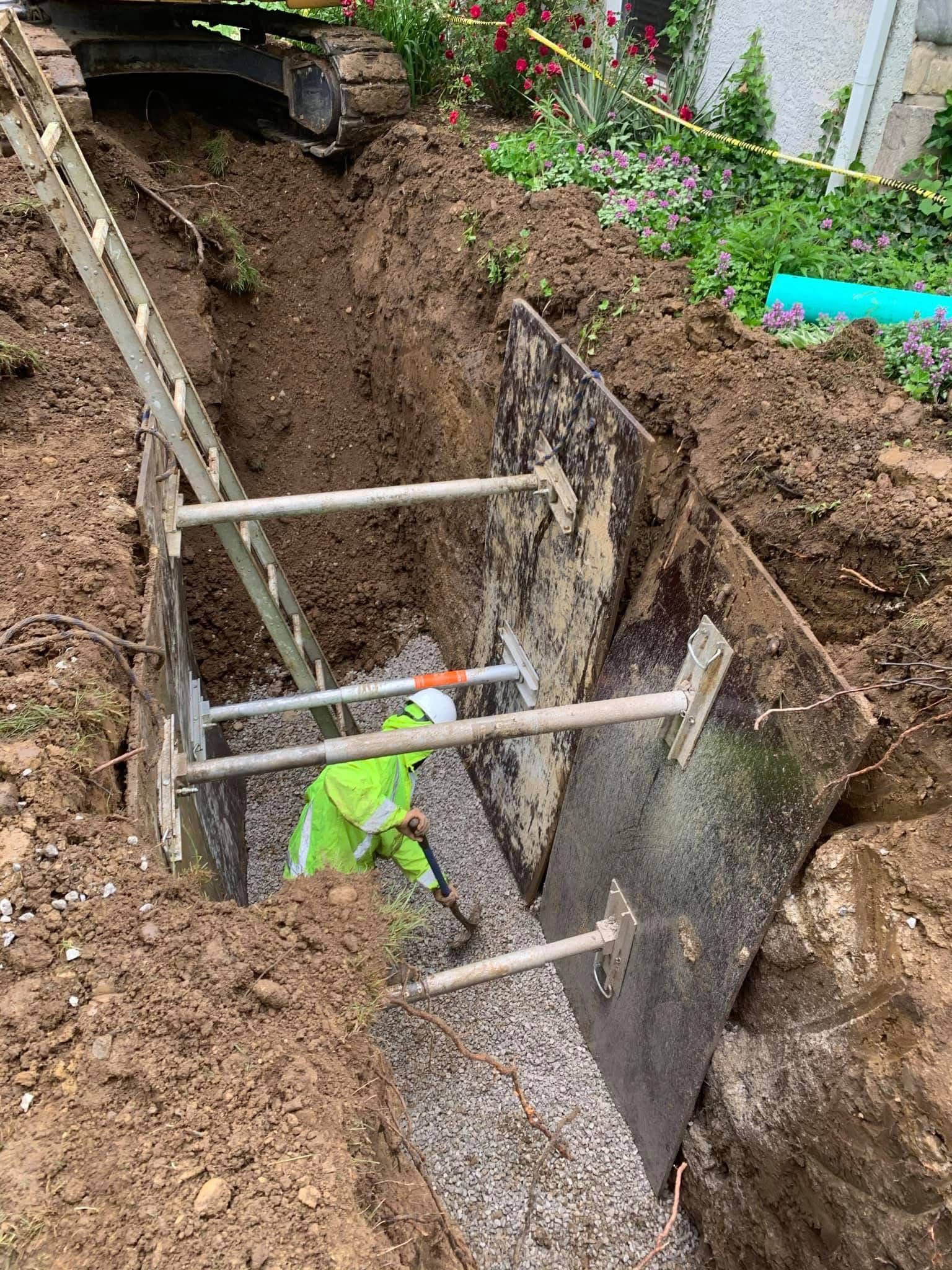 Worker in neon vest inside a deep trench with bracing boards during excavation work.