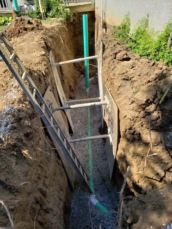 Trench with gravel base and green pipe under construction, flanked by dirt walls and wooden supports