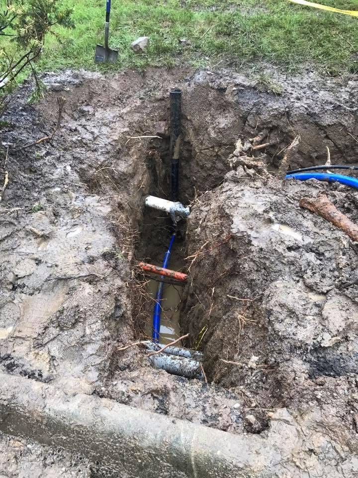 Trench in muddy ground exposing a blue pipe with white and orange fittings.