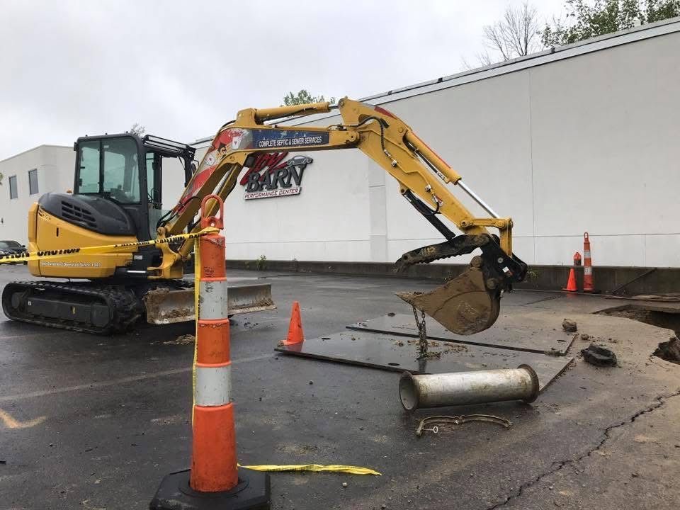 Yellow excavator on wet pavement beside a white building, with orange cones and a fallen barrel nearby.