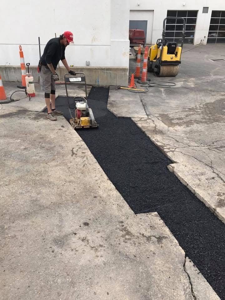 Worker paving a black asphalt strip beside a warehouse, using a compacting machine.