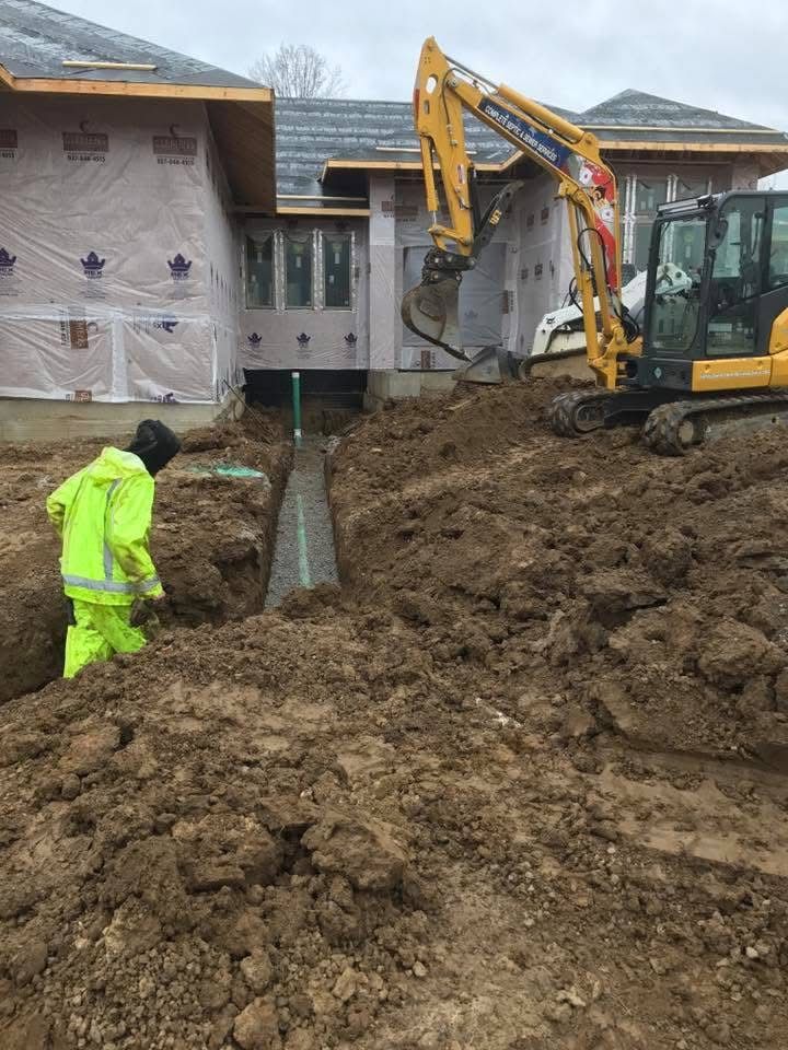 Construction workers digging a trench beside a house, with an excavator and muddy soil.