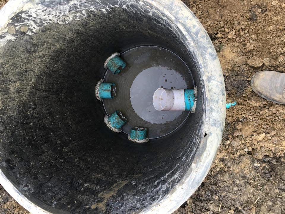Open manhole with a gray pipe and blue fittings inside, surrounded by dirt.