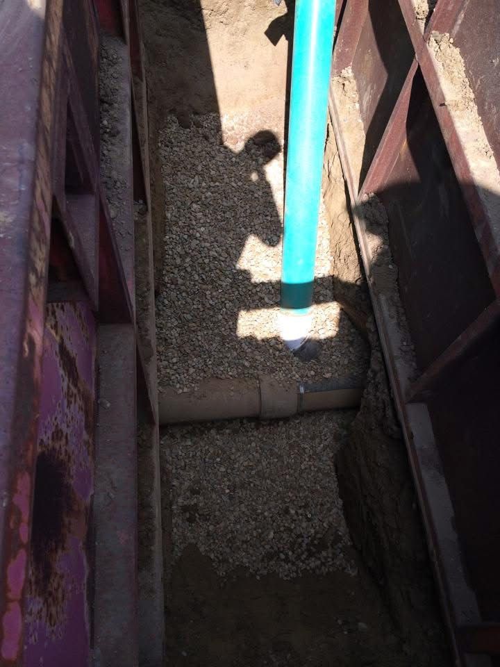 Blue hose spraying water into a narrow trench filled with gravel and dirt between metal walls.
