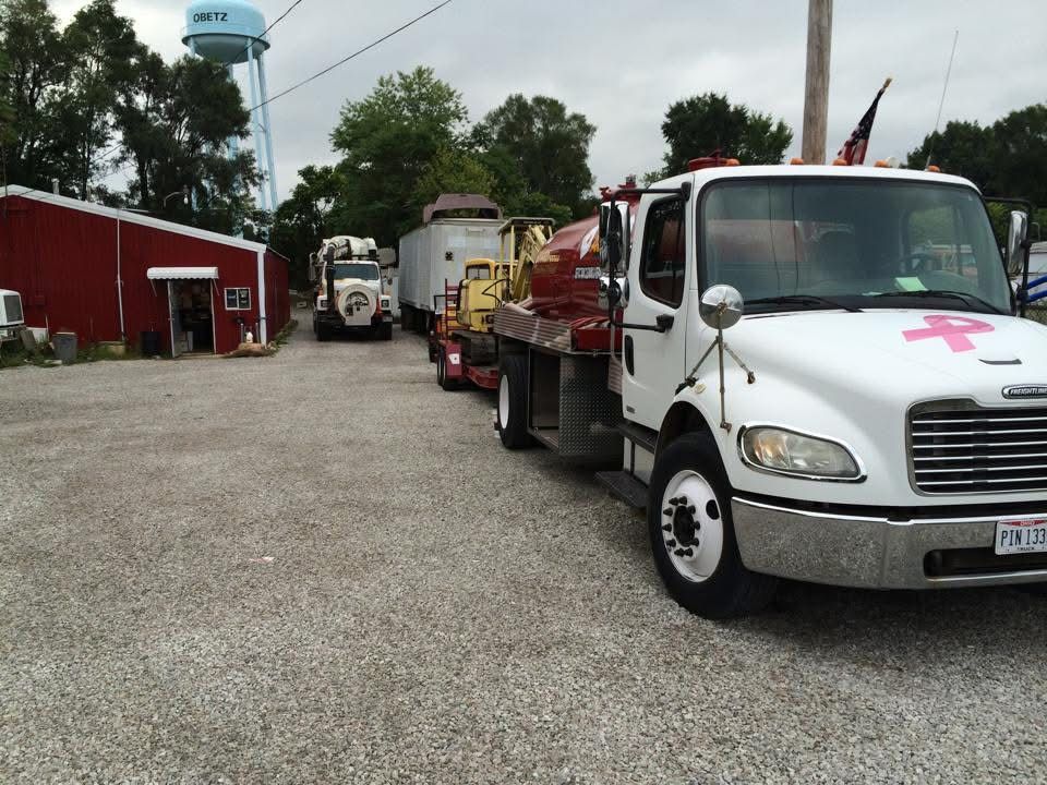 White tow truck with pink ribbon emblem parked beside other vehicles on a gravel lot near a red building.