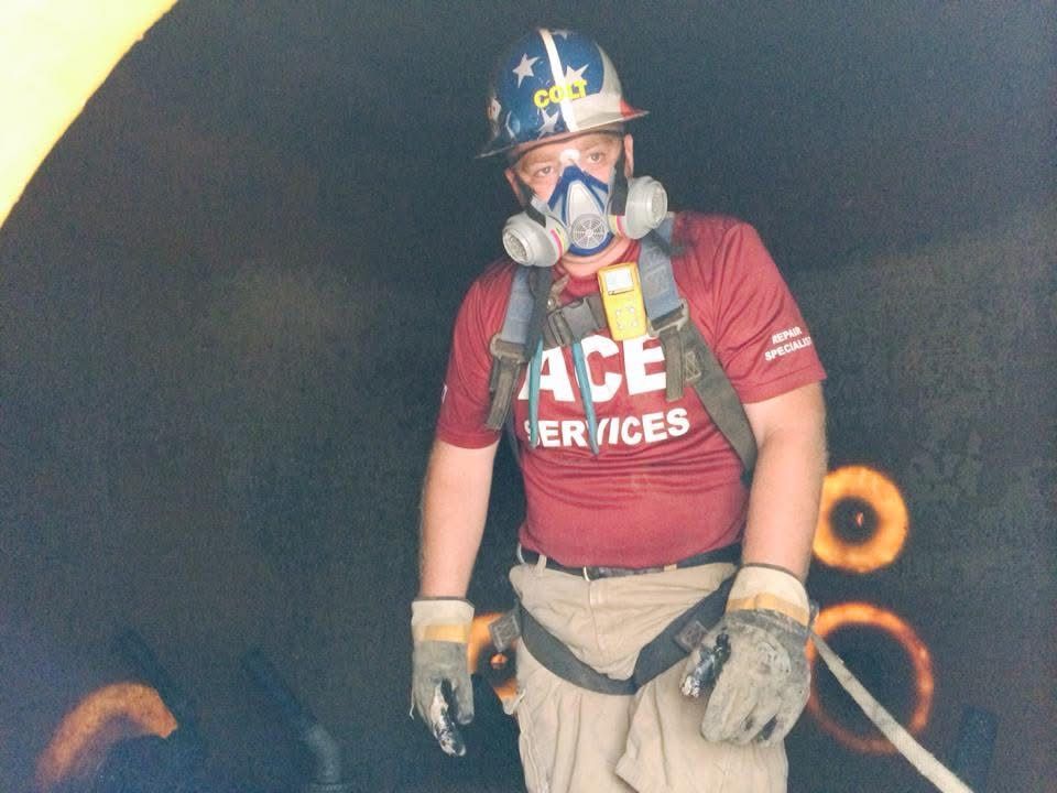 Worker in helmet and respirator, wearing a red ACE Services shirt and safety gear in a smoky area.