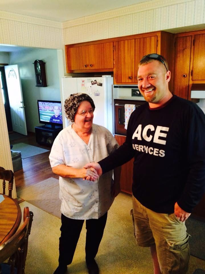 Two people smiling in a kitchen, one holding a tool and wearing a black ACE Services shirt.