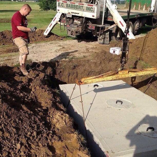 Worker guiding a crane lowering a concrete septic tank into a dirt excavation site