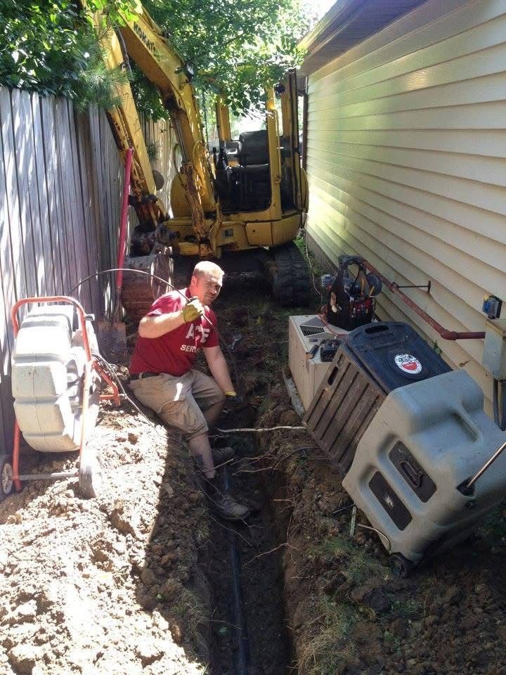Worker in red shirt stands in a narrow trench beside a house, with an excavator and debris nearby.