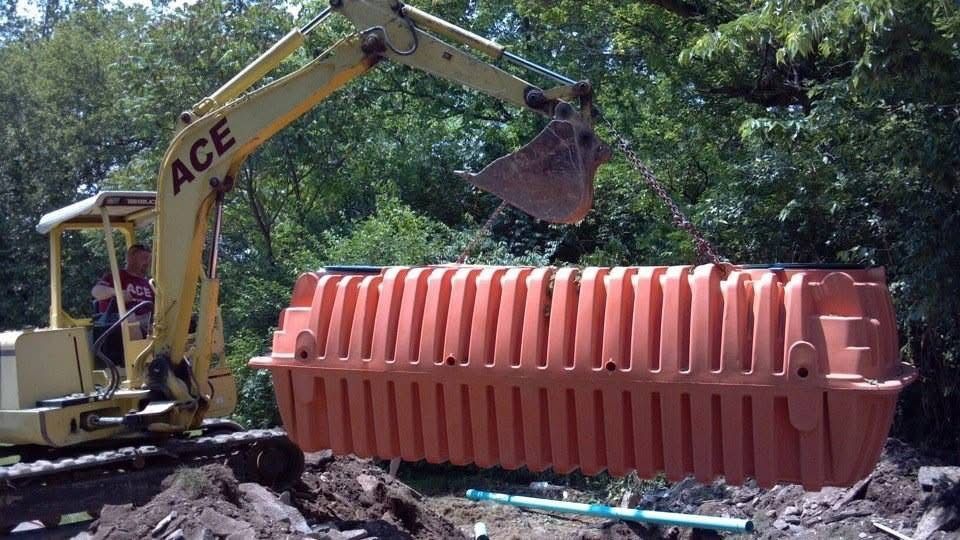 Excavator loading a large orange plastic tank at a construction site surrounded by trees