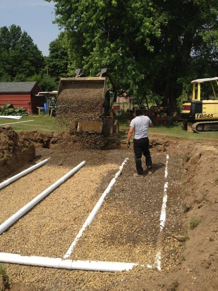 Worker at a construction site laying white drainage pipes in an excavated trench beside a yellow excavator.