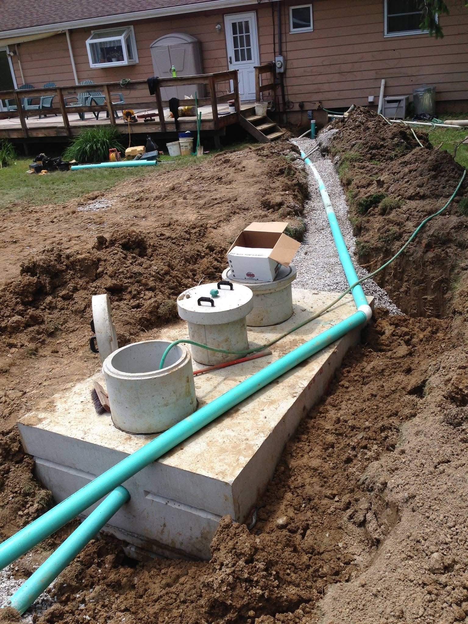 Outdoor utility installation beside a house, with PVC pipes and concrete boxes in a muddy trench.