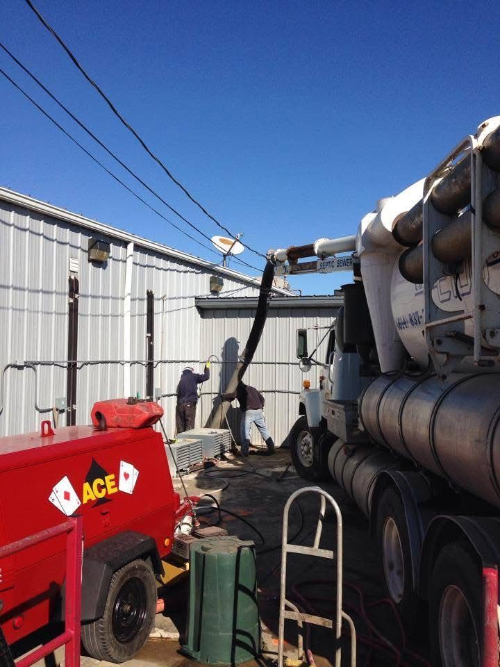 Construction yard with red ACE trailer, large tank truck, and utility equipment between metal buildings.