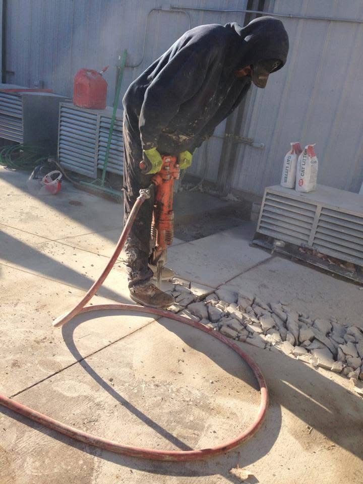 Worker using a jackhammer to break concrete on a sidewalk, with a red hose in the foreground