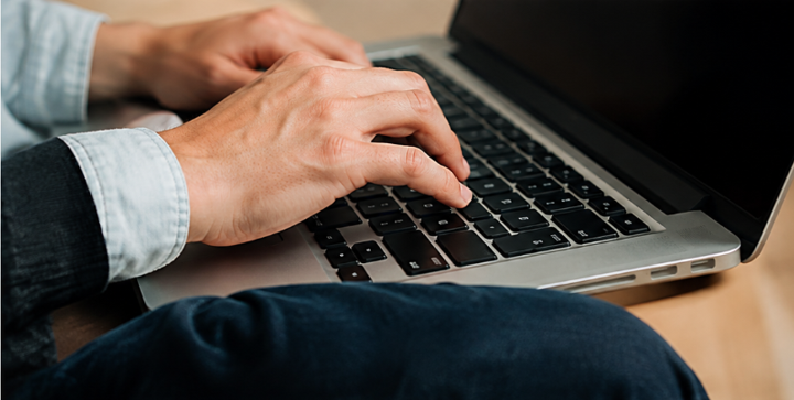 Hands typing on a laptop keyboard beside a black screen on a wooden table.